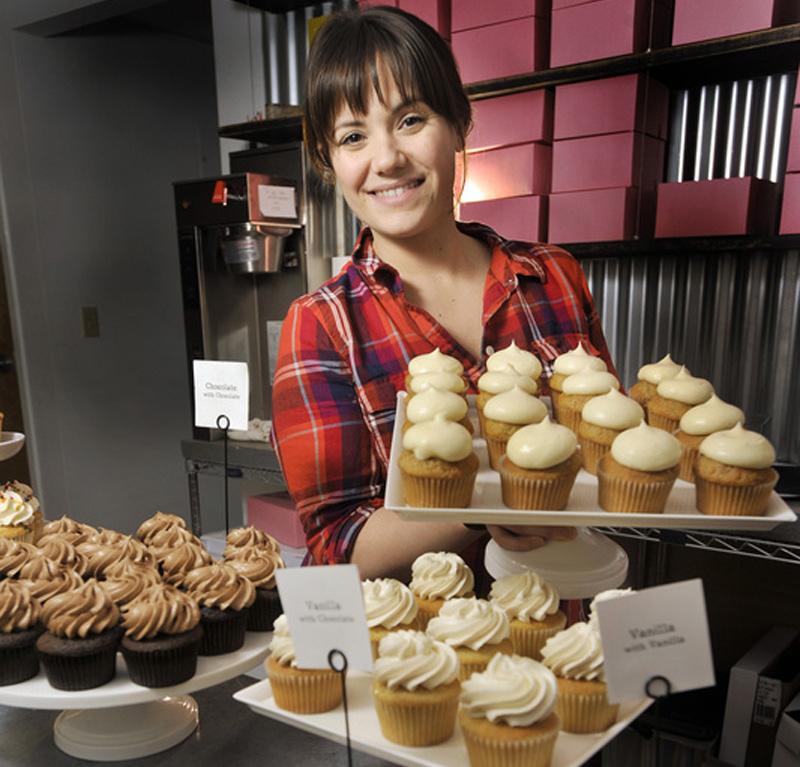 Sarah Crocker is wearing an orange, red, and purple plaid shirt and holding a tray of white cupakes frosted in white icing. She has brown hair and is in a room surrounded by other cupcakes and cake boxes.