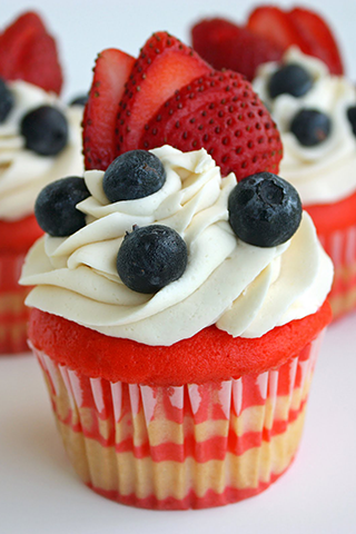 Red and white cupcake frosted in white and covered in bluberries and sliced strawberries